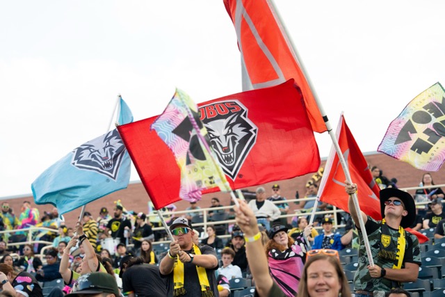 An array of flags being waved in the supporter's section at a NM United game. Meow Wolf and UNM flags are visible.