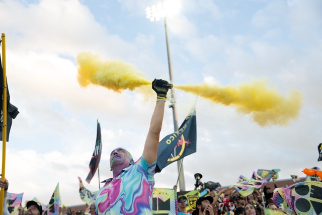 A man painted in many colors hold up a yellow smoke bomb in front of the supporter's section.