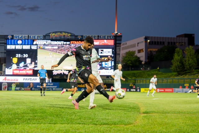 A NM United player kicks the ball while the sun sets.
