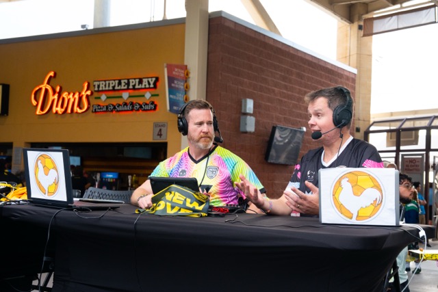 Two podcast hosts converse in front of a Dion's pizza on the concourse at a NM United game.