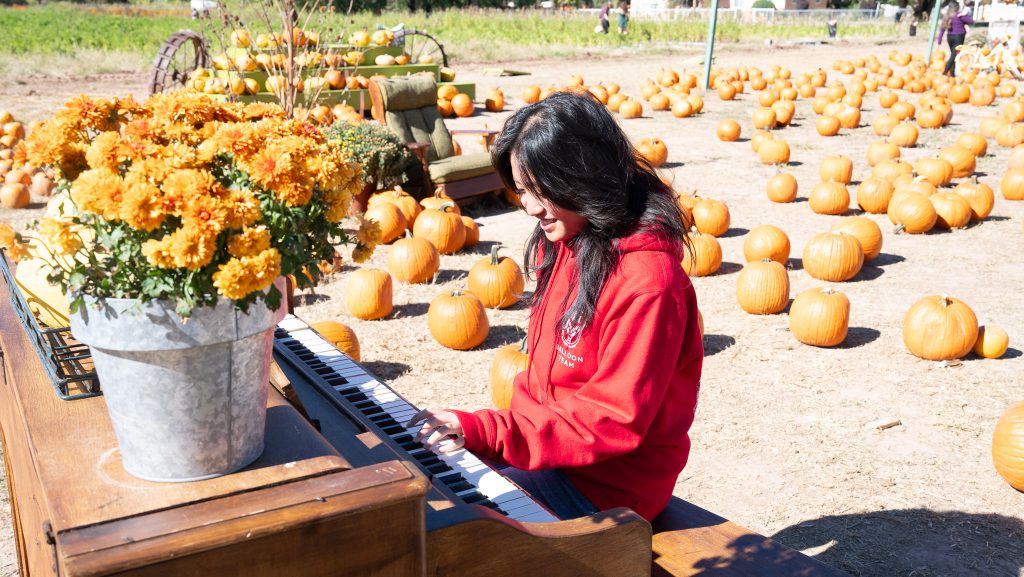 A student smiles as she plays an old piano outside with pumpkins seen on the ground.