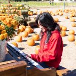 A student smiles as she plays an old piano outside with pumpkins seen on the ground.