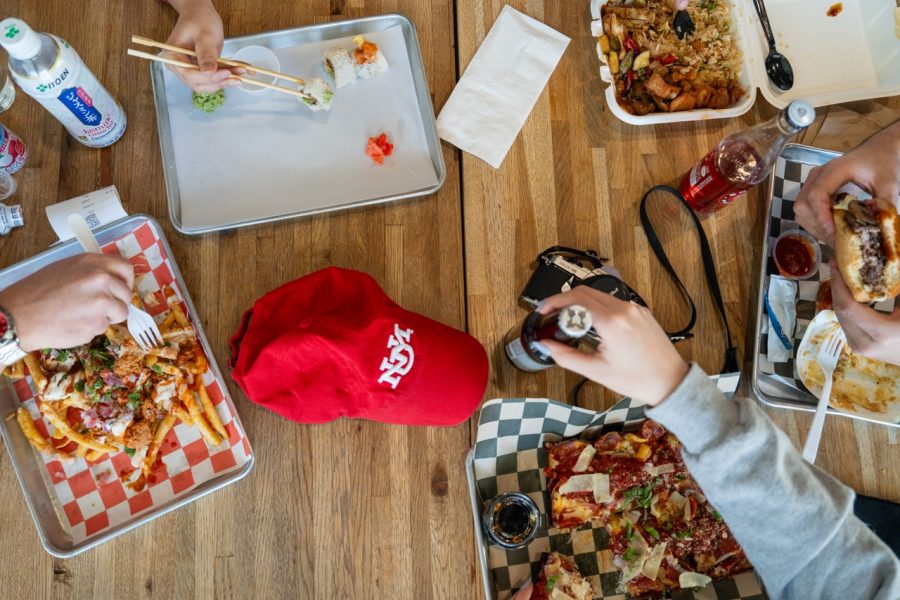 An overhead shot shows a table layout with plates of food, bottles of drinks, and a cherry red UNM hat.