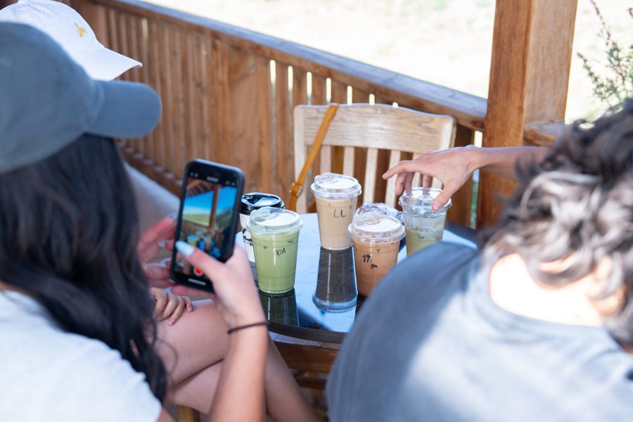 A person takes photos of their drinks on an iPhone on an outdoor patio with wooden furniture. There are two iced coffees, one iced matcha, one hot matcha and one iced tea.