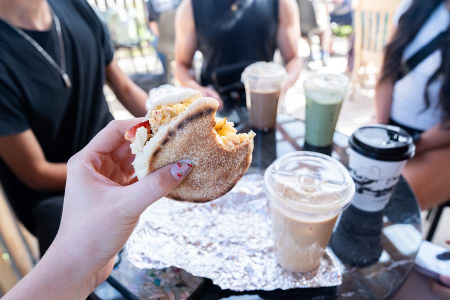 A person holds a breakfast sandwich up to the camera with drinks on a table visible in the background.