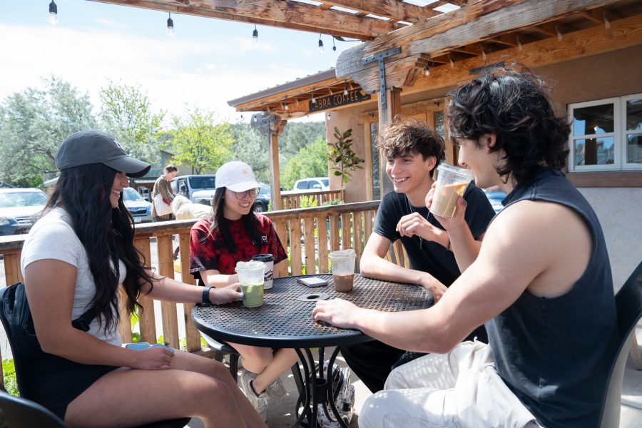 Four students in UNM merchandise smile while drinking their drinks on an outdoor wooden patio. In the background, you can see trees and a mountainscape.