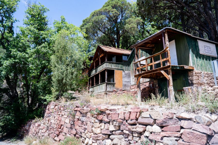 Old wooden structures sit on top of a hill with a stone wall, surrounded by lush trees.