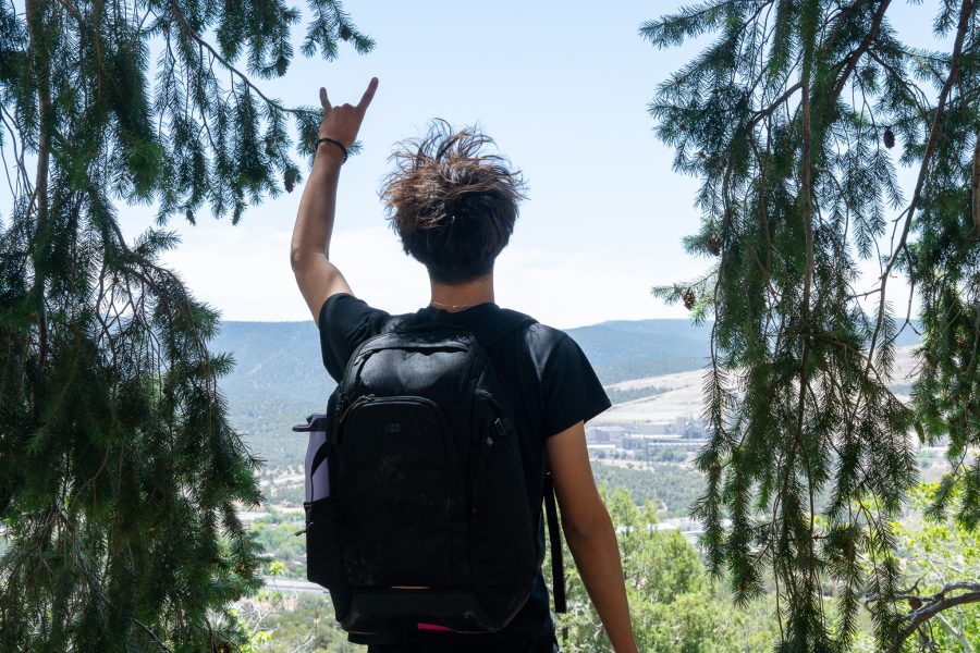 A UNM student stands at the top of the Carlito Springs Trail, holding up a Lobo hand symbol, and looking down towards Cedar Crest.