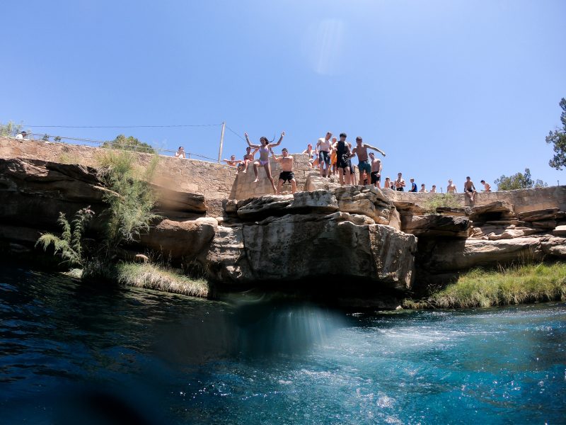 A group of individuals are jumping from a rock ledge into the clear blue water of Blue Hole, surrounded by stone formations and greenery. A few onlookers stand nearby.