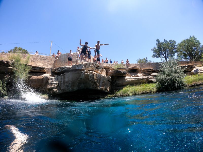 A group of individuals are jumping from a rock ledge into the clear blue water of Blue Hole, surrounded by stone formations and greenery. A few onlookers stand nearby.