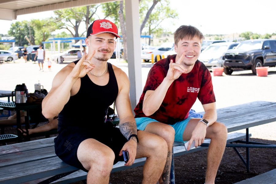 Two students wearing UNM merchandise hold up a Lobo hand symbol. They sit in a shaded structure at a picnic table.