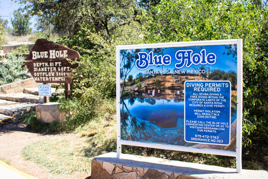 Two signs for the Blue Hole are shown with trees in the background on a sunny day.